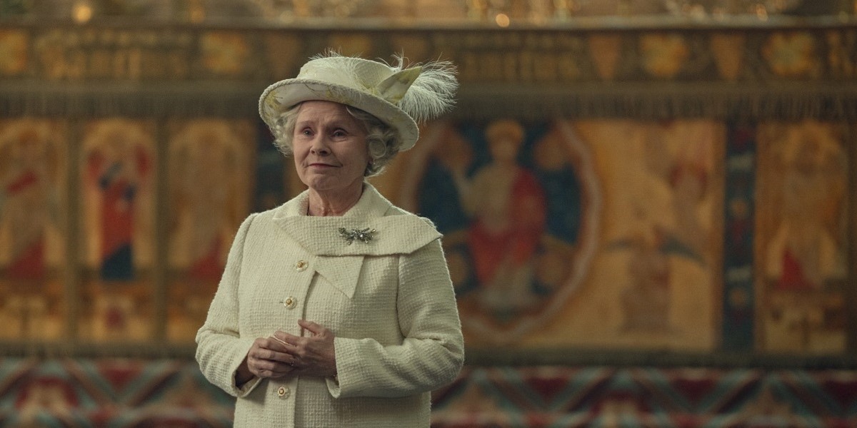 Image of Imelda Staunton as Queen Elizabeth II in Netflix's 'The Crown." She is a white elderly woman with short, curled white hair standing in a church with a tapestried wall behind her. She's wearing a white hat with a feather in it and a white coat with a diamond pin on it. She's fiddling with her wedding ring as she looks into the distance with a melancholy smile on her face. 