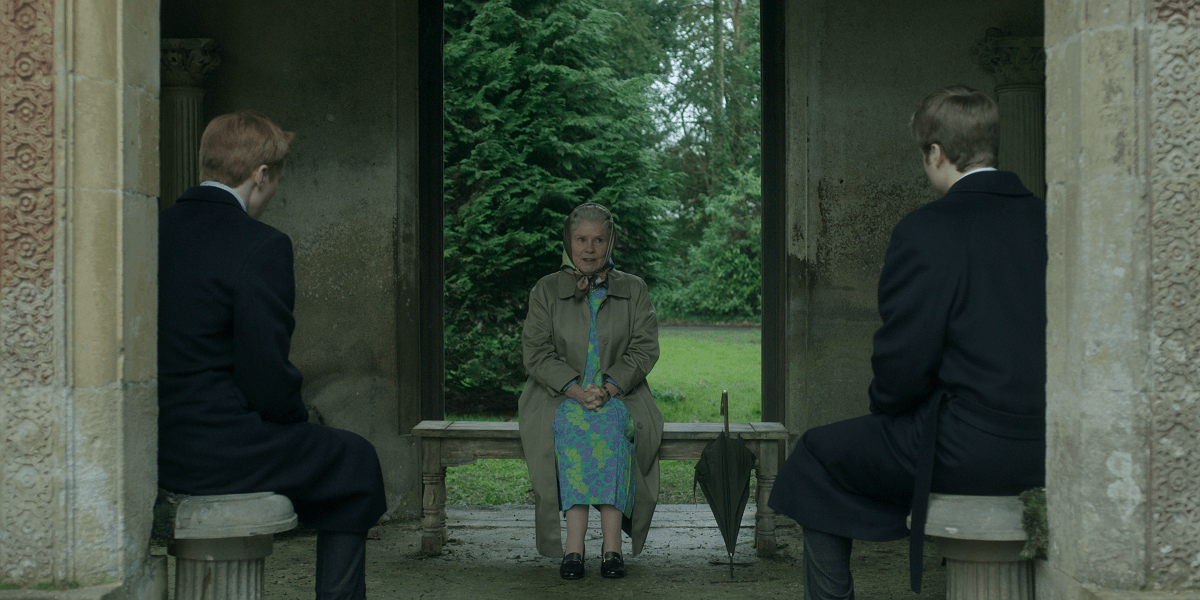 Image of Luther Ford as Prince Harry, Imelda Staunton as Queen Elizabeth II, and Ed McVey as Prince William in a scene from Netflix's 'The Crown." They are seated in a stone gazebo. Harry and William are white teenage boys in black suits with their backs to the camera. Elizabeth is an elderly white woman seated between them on a stone bench facing the camera. She's wearing a kerchief around her white hair and a trench coat over her blue, purple, and green flower print dress. An umbrella leans on the bench beside her. 