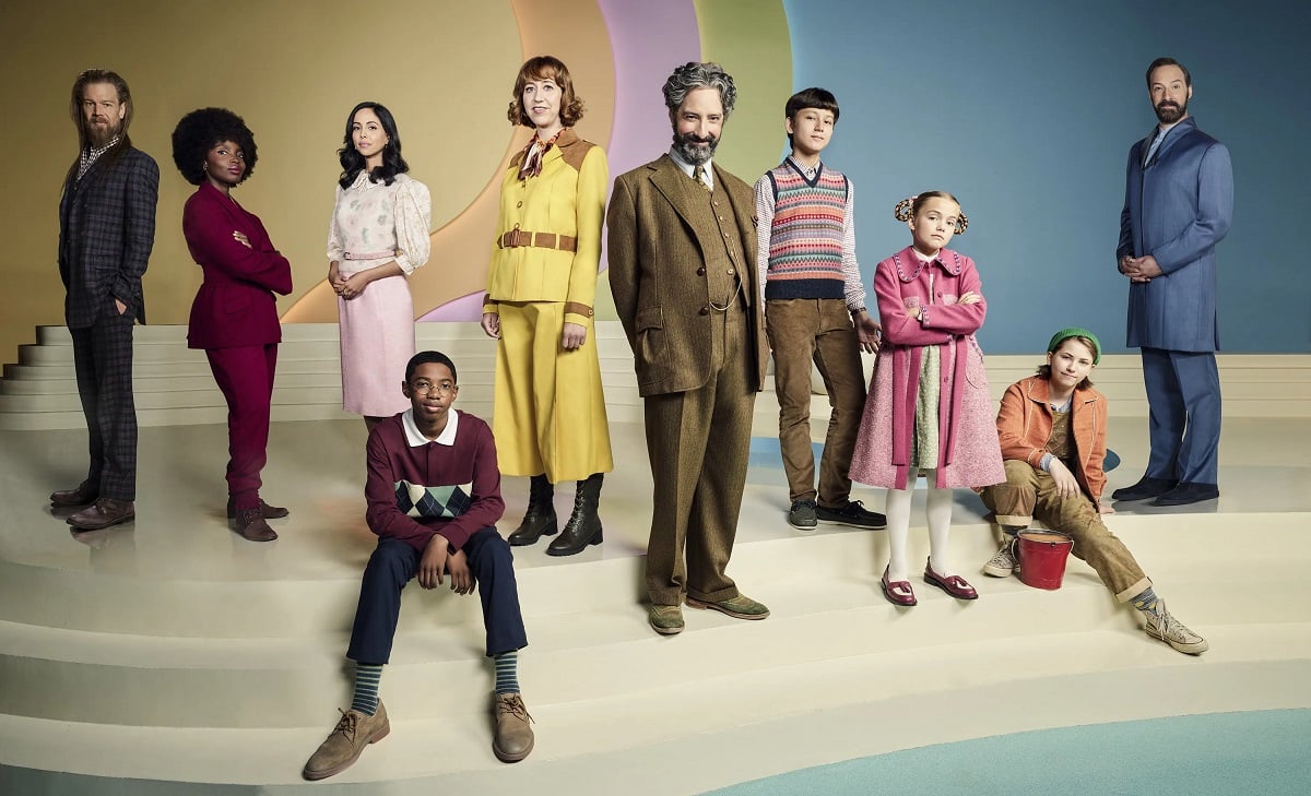 A promotional image of the cast of 'The Mysterious Benedict Society.' A mixed group of 10 people (6 adults and 4 children) stand or sit on a beige, stair-like platform in front of a pastel-colored background. They are all in costume as their characters.