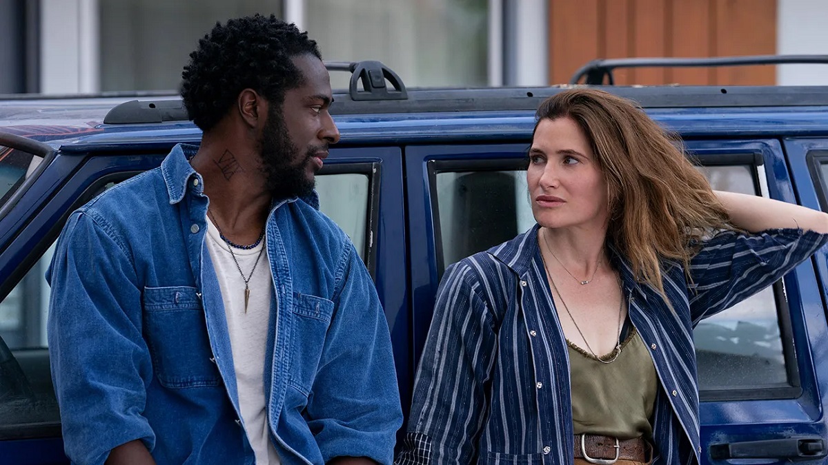 Image of Quentin Plair and Kathryn Hahn in a scene from 'Tiny Beautiful Things.' Plair is a Black man with short black hair in twists and a beard. He's wearing a denim buttondown shirt over a white t-shirt and has a tattoo on his neck. Hahn is a white woman with long brown hair wearing a blue and white striped buttondown open over a green silk blouse. They are looking at each other and both leaning against a blue SUV. 