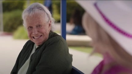 An old woman (Ann Roth) sits on a bench smiling at Barbie (Margot Robbie).