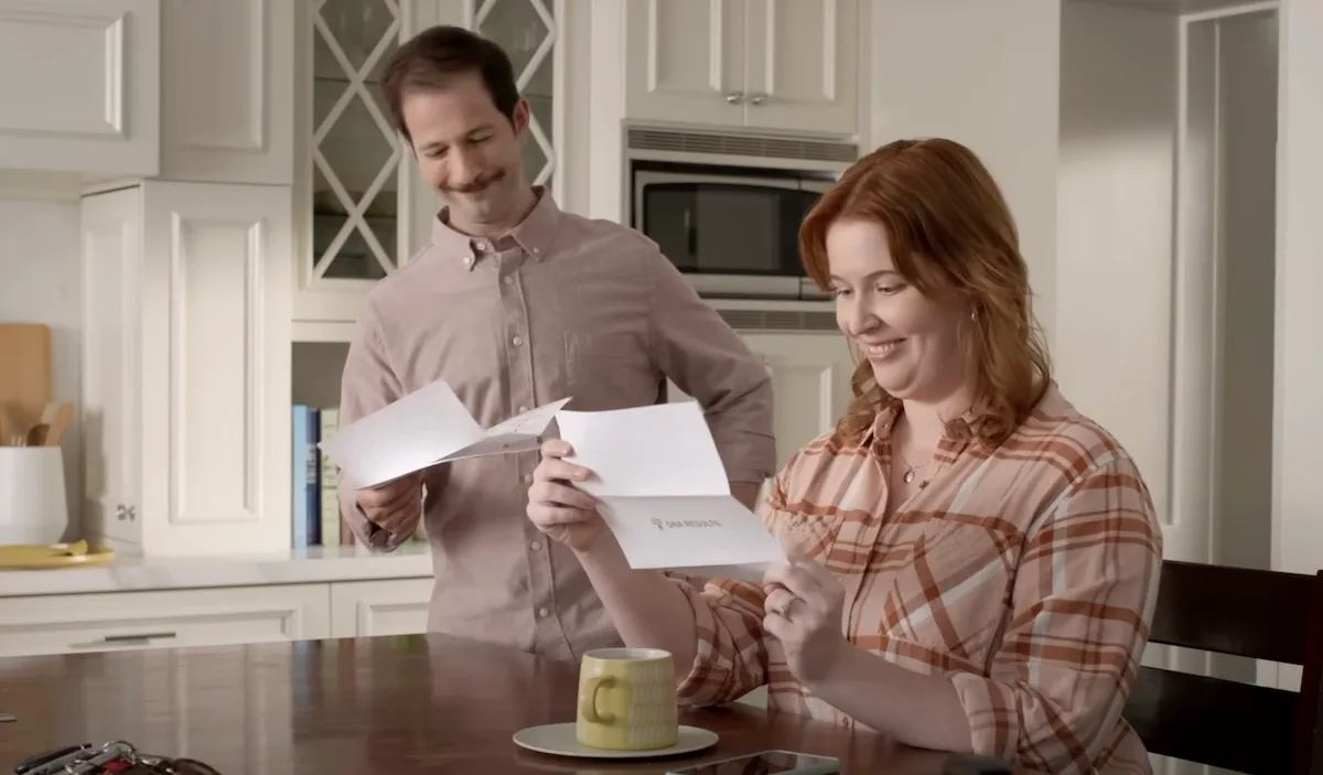 A man and woman sit in their kitchen, smiling over letters that say "DNA results."