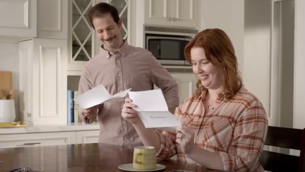 A man and woman sit in their kitchen, smiling over letters that say 