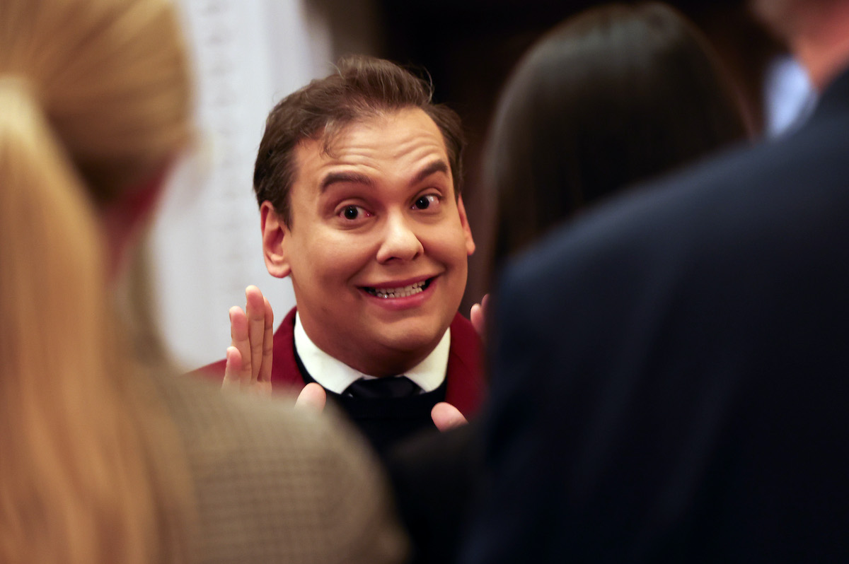 George Santos smiling, seen between the shoulders of two unidentifiable people.