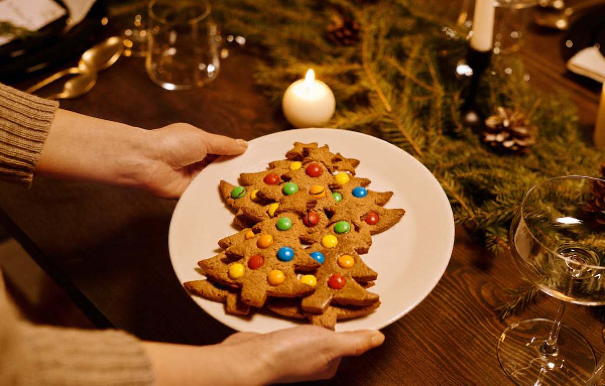 Two hands set a plate of Christmas tree-shaped cookies onto a table decorated with greenery.