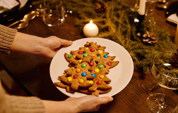 Two hands set a plate of Christmas tree-shaped cookies onto a table decorated with greenery.