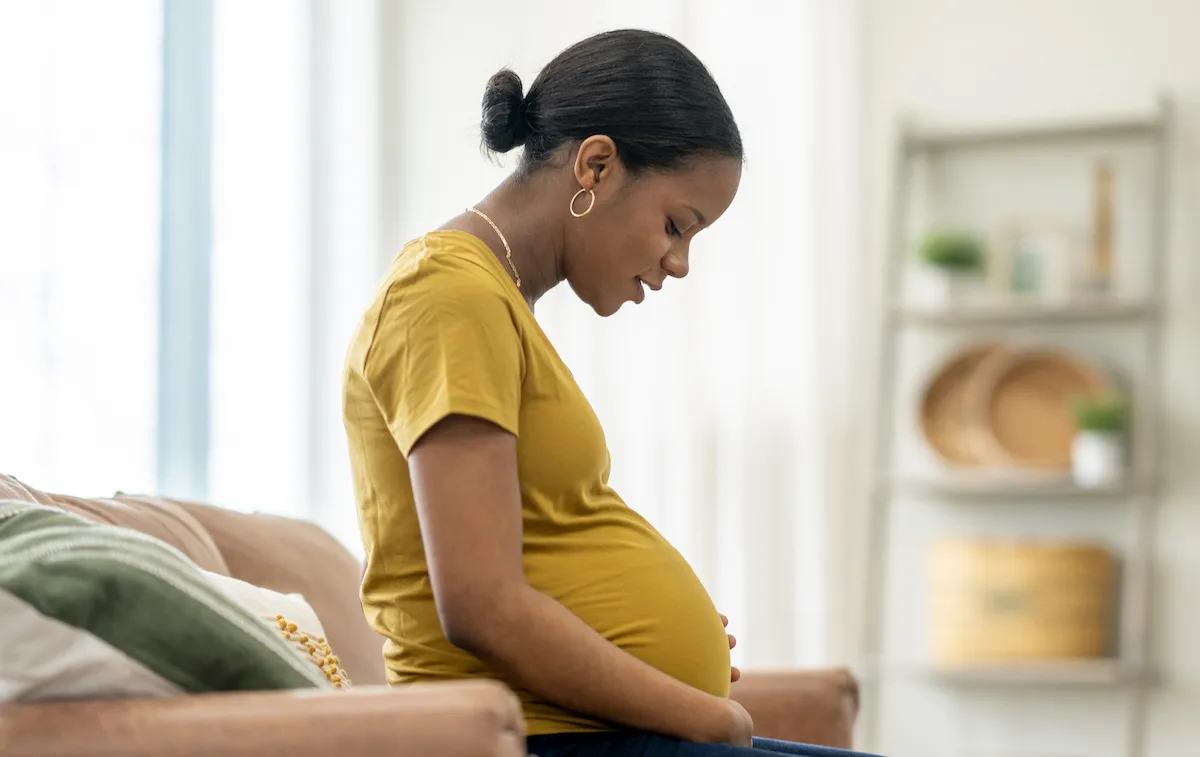 A young pregnant woman of African decent sits on a sofa in the comfort of her own home as she cradles her belly with her hands. She is dressed casually as she looks down at her belly with anticipation.