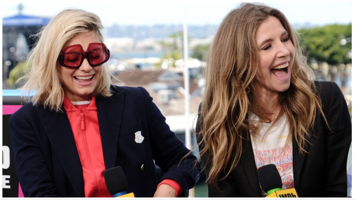 Spencer Grammar in pink sunglasses and a black blazer and sarah chalke in a white shirt and black blazer laugh.