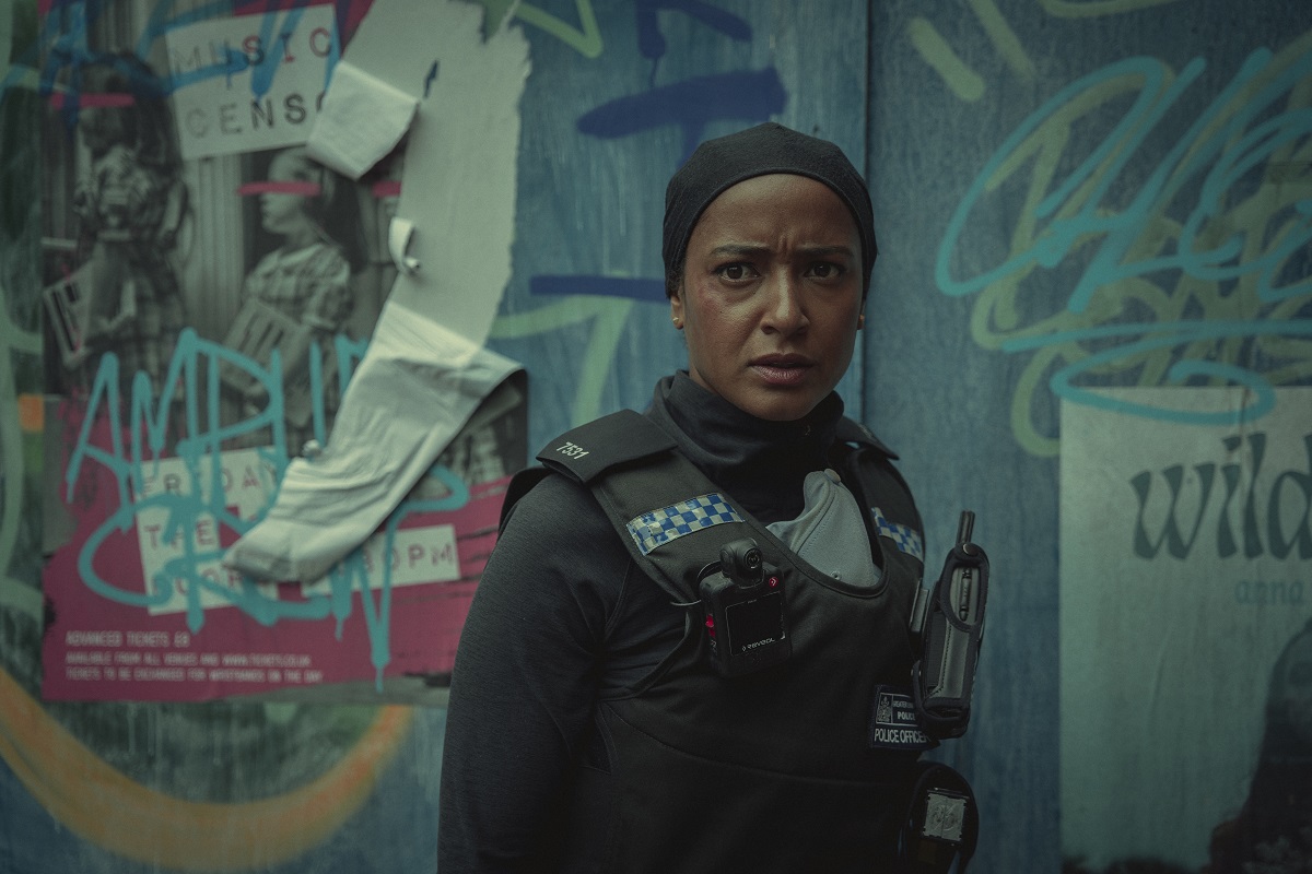 Image of Amaka Okafor as DS Hasan in Netflix's 'Bodies.' She is a Black Muslim woman with her hair covered and wearing a black police uniform. She is standing in front of a blue, graffiti covered wall.