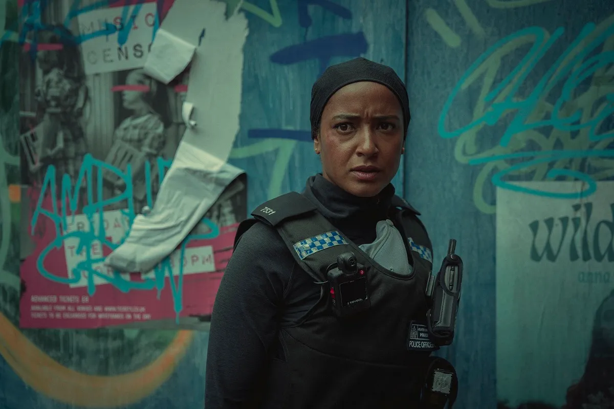 Image of Amaka Okafor as DS Hasan in Netflix's 'Bodies.' She is a Black Muslim woman with her hair covered and wearing a black police uniform. She is standing in front of a blue, graffiti covered wall.