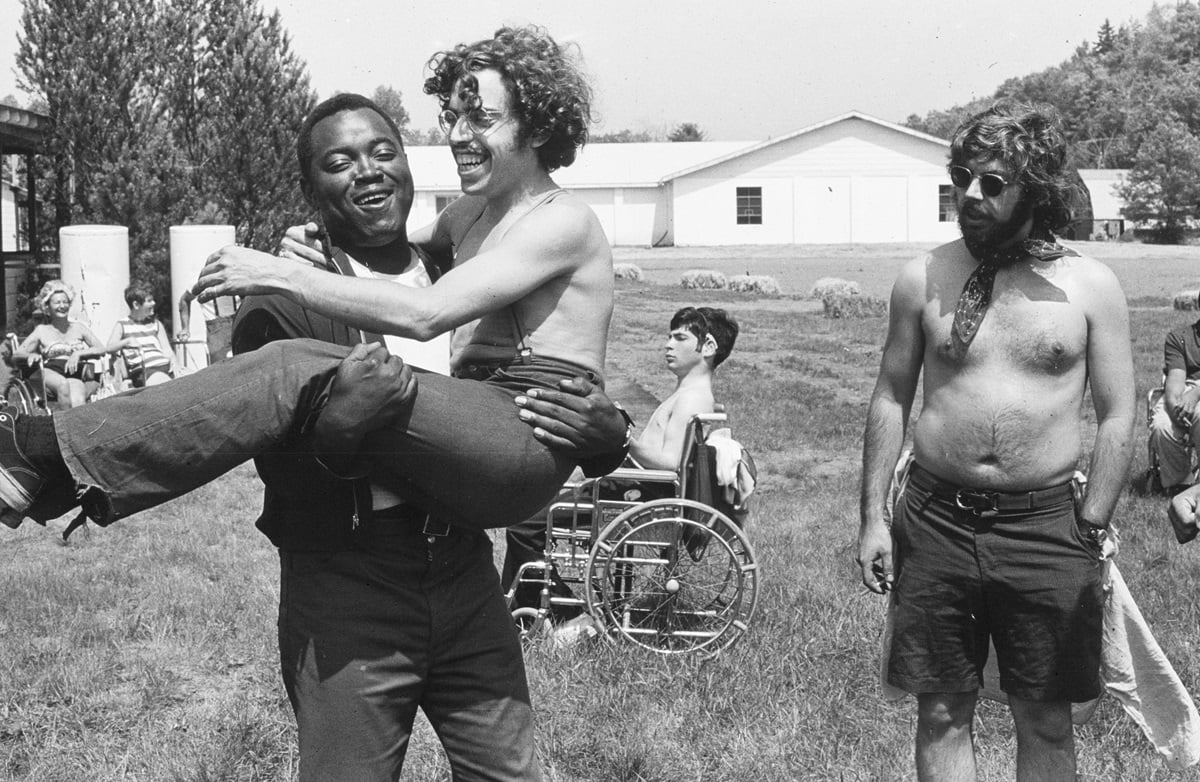A man holds another man aloft while a man in a wheelchair sits behind them in the field