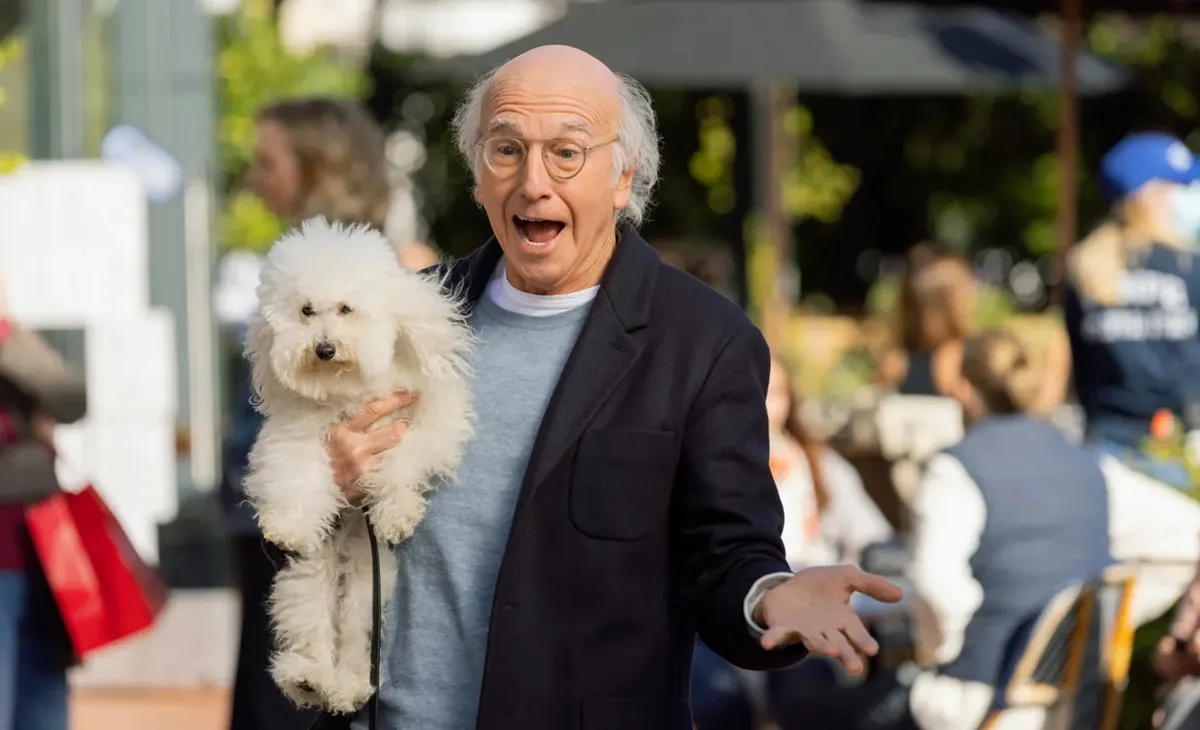 Larry David looks surprised while holding a small white dog in Curb Your Enthusiasm