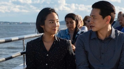 Greta Lee and Teo Yoo sit on a bench looking at one another with the ocean behind them