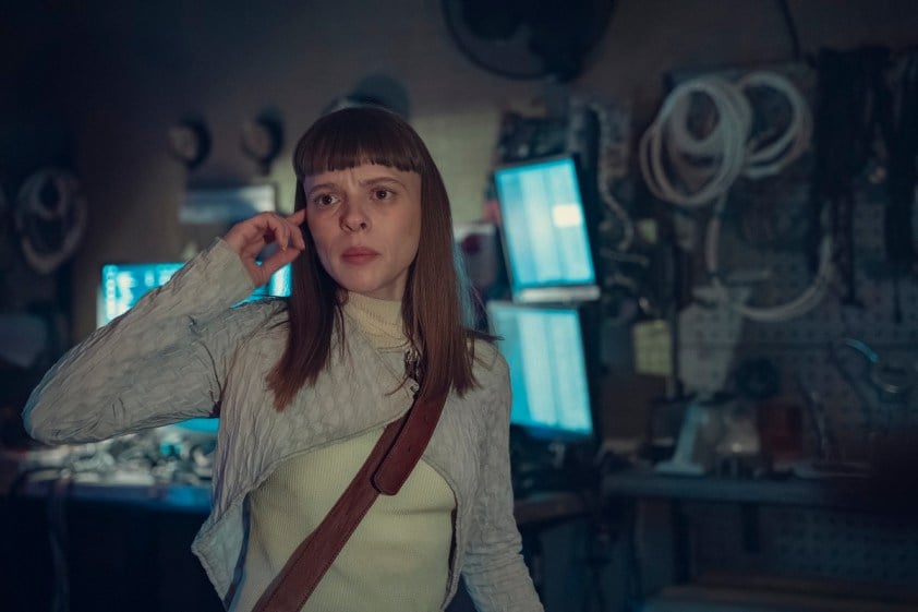 Shira Haas as Iris Maplewood in a scene from Netflix's 'Bodies.' She is a white woman with long light brown hair and bangs. She's wearing an asymmetrical grey knit cardigan over a white turtleneck with a brown crossbody bag strap across her chest. She's standing in a room with several computer screens and tools in it. 