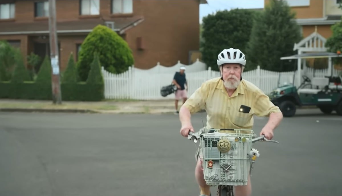 An older white man rides a bicycle with a basket through a suburban neighborhood.