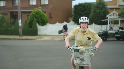 An older white man rides a bicycle with a basket through a suburban neighborhood.