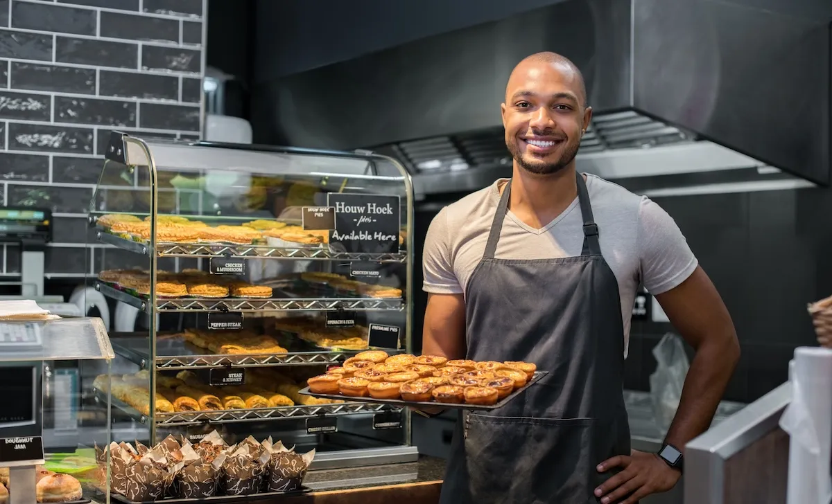 Smiling Black baker with apron holding tray of small pastry and looking at camera.