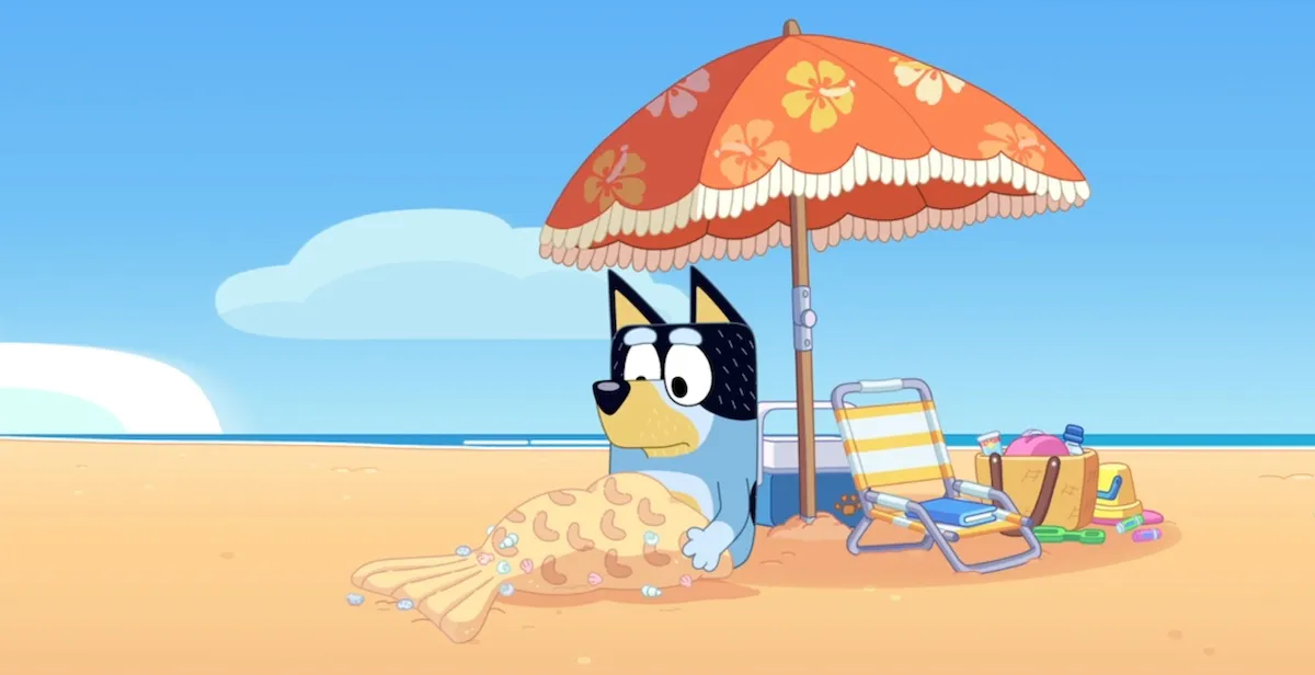 Bandit sits glumly under a beach umbrella, with his legs covered in a mermaid tail made of sand.