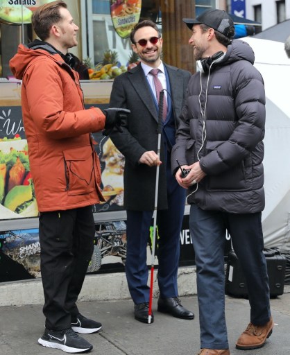 Charlie Cox, dressed as Matt Murdock, speaks to two men in jackets on a sidewalk.