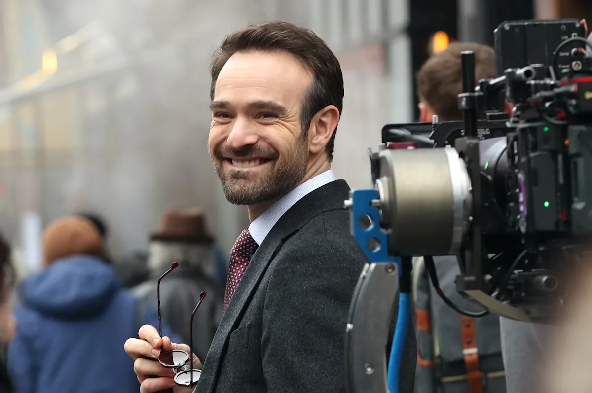 Charlie Cox smiles, holding his sunglasses in costume as Matt Murdock. A large camera is visible on the right.
