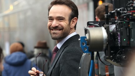 Charlie Cox smiles, holding his sunglasses in costume as Matt Murdock. A large camera is visible on the right.
