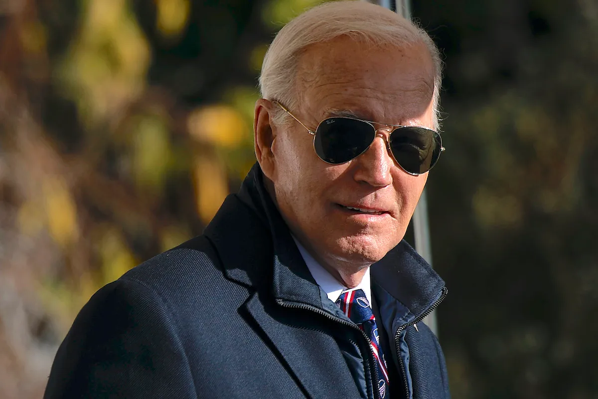 Joe Biden gives a slight smile to the camera, wearing a coat and aviator sunglasses.
