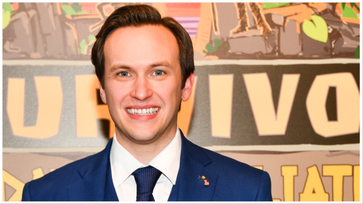 Nick Wilson smiles awkwardly while wearing a blue suit.