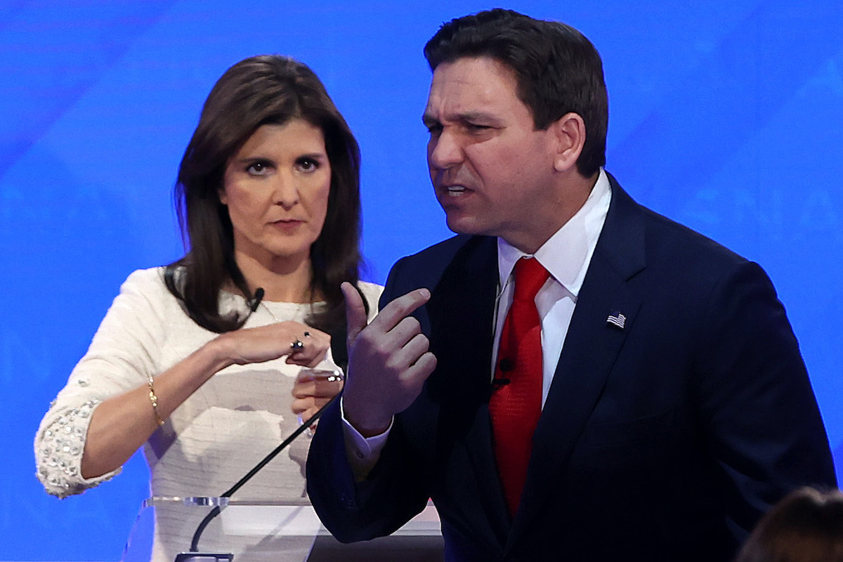 Ron DeSantis checks his teeth and Nikki Haley stands behind him glaring at him during a break at a Republican debate.