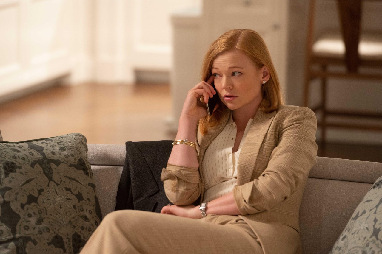 Sarah Snook as Shiv Roy, sitting on a couch in a tan suit, holding a cell phone to her ear in 'Succession'.