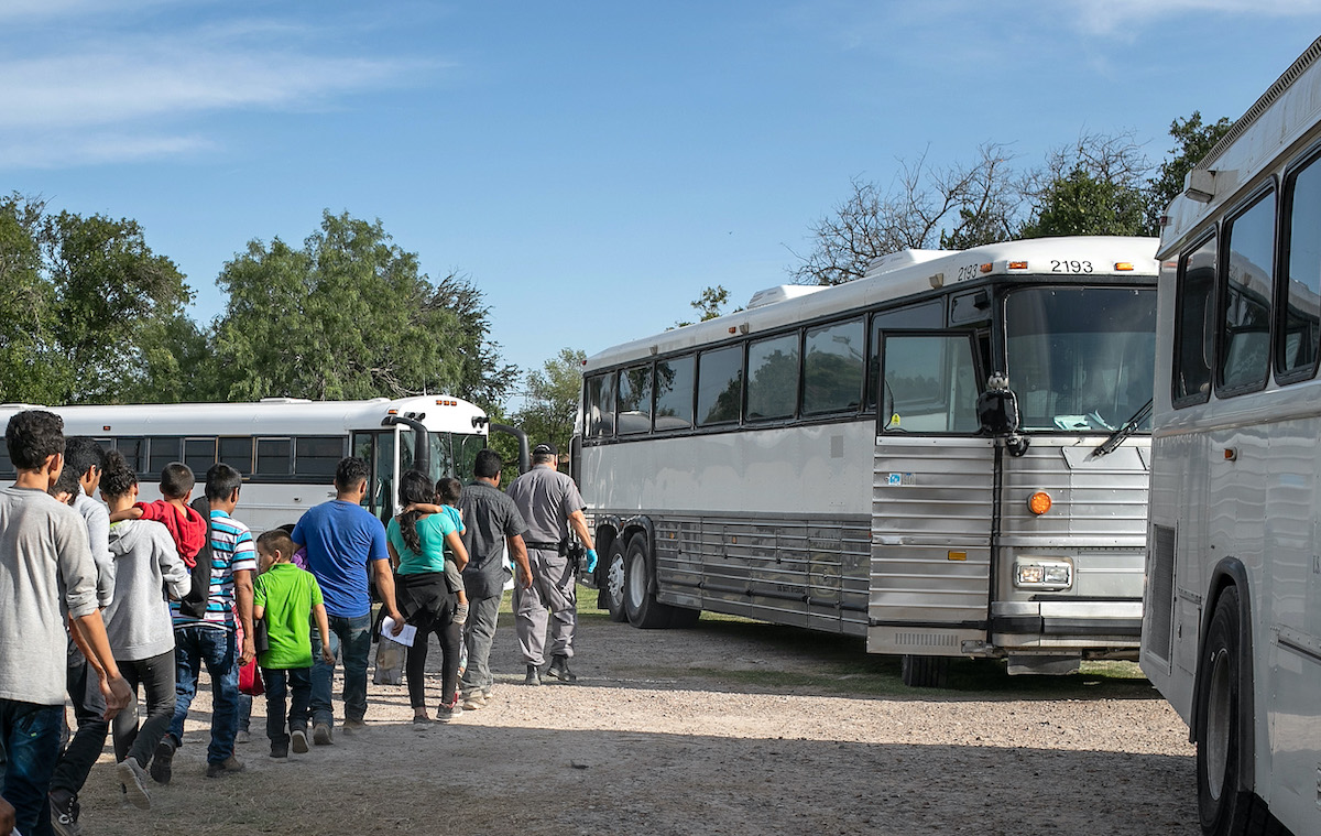 A line of migrants boards a large white bus.