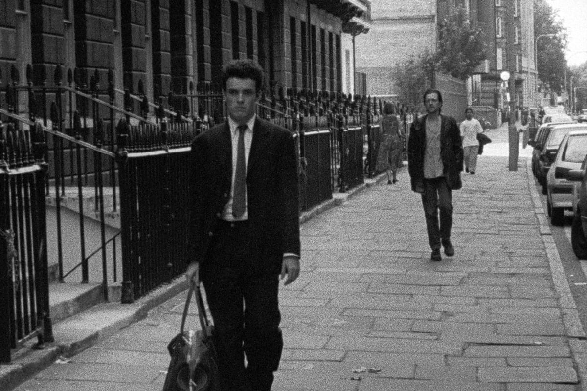 Black and white image of Alex Haw as Cobb and Jeremy Theobald as Bill in a scene from 'Following.' Cobb is a young white man with short, dark hair wearing a suit and tie and carrying a large tote bag down a street in London. Bill is a young white man with shaggier brown hair wearing a leather jacket over a buttondown and jeans following Cobb at a distance.