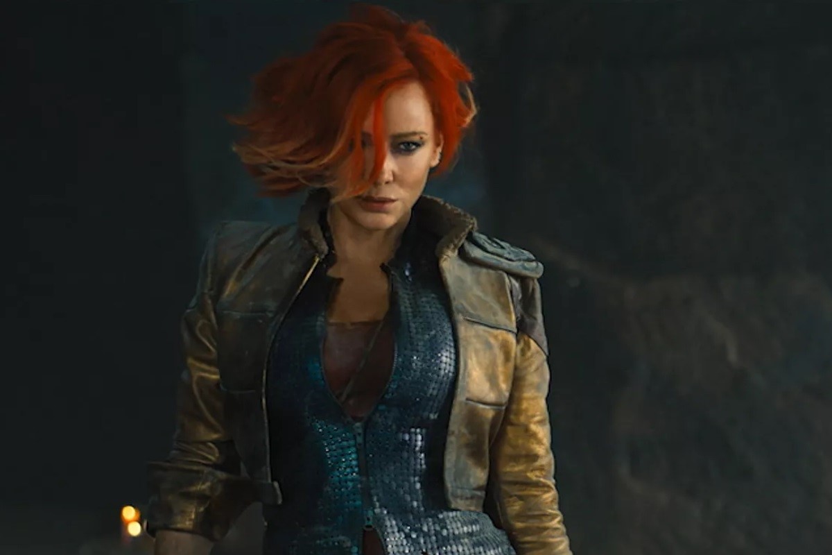 Cate Blanchett as Lilith in a scene from 'Borderlands.' She is a white woman with bright red, swooping, chin-length hair wearing a brown jacket layered over a black, armored zip-up shirt, over a tank top. 