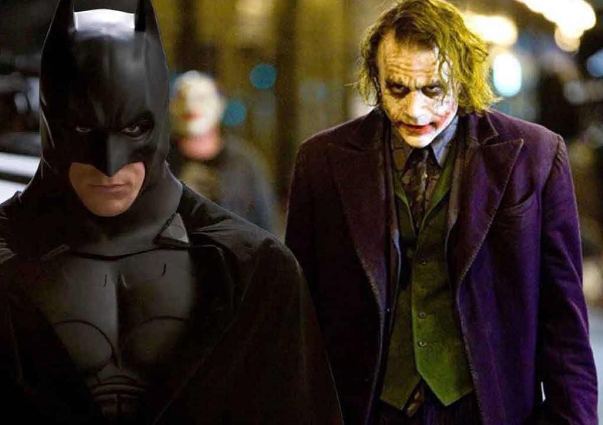 Christian Bale as Batman and Heath Ledger as The Joker in a scene from 'The Dark Knight." Batman is a white man in a black Bat suit that covers him entirely except for his mouth and chin. The Joker is a white man in messed up clown make-up with medium length, stringy hair that's been dyed green, and wearing a purple blazer over a green vest, a blue buttondown and a patterned tie. Batman is walking forward and The Joker is following closely behind.