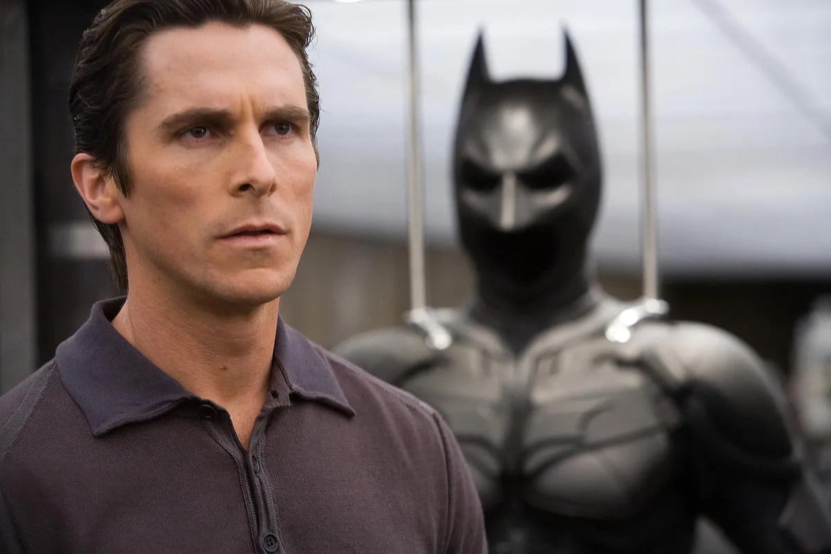 Christian Bale as Bruce Wayne in a scene from 'Batman Begins.' Wayne is a white man with short dark hair wearing a purple-grey polo shirt. He's standing in front of his Batman suit.