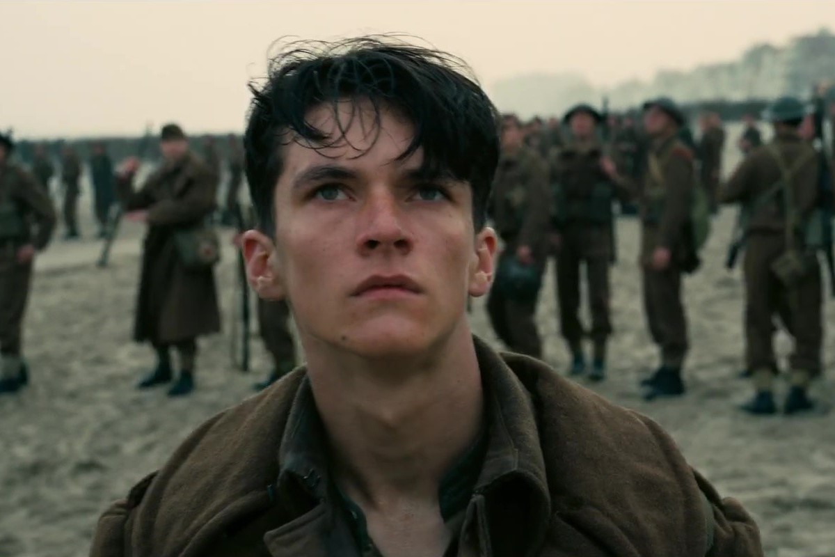 Fionn Whitehead as Tommy in a scene from 'Dunkirk.' Tommy is a young white man with shaggy dark hair wearing a 1940s British army uniform. He stands on a beach looking up with other soldiers in the background behind him. 
