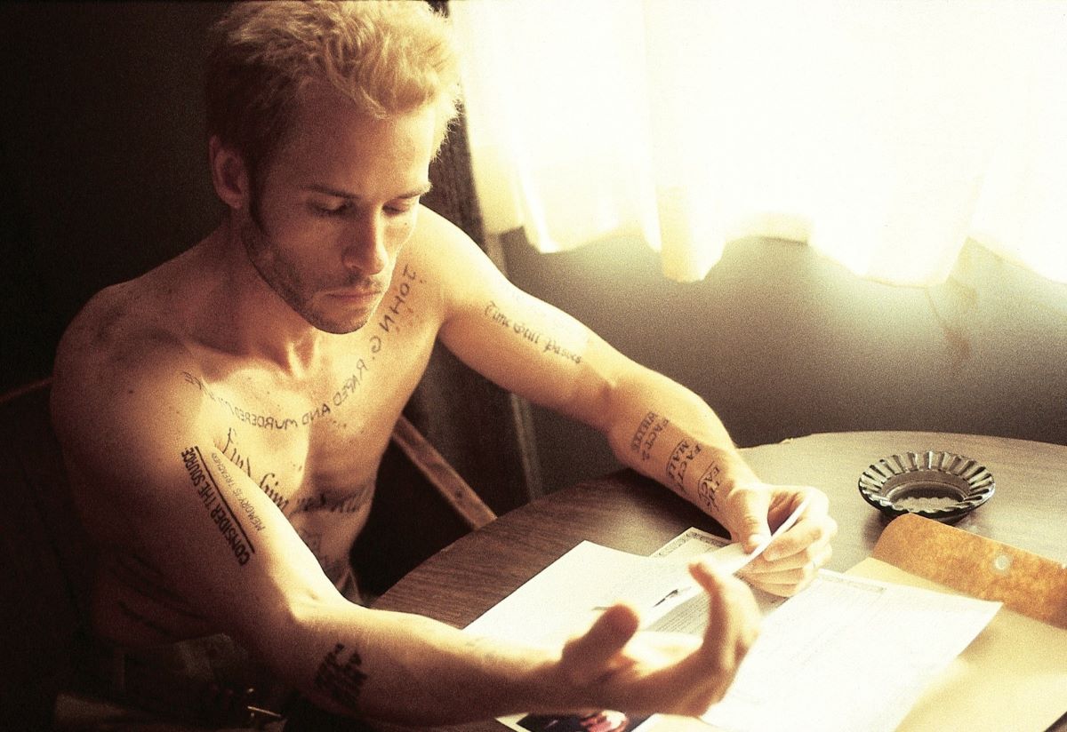 Guy Pearce as Leonard in a scene from 'Memento.' Leonard is a white man with short, frosted blond hair. He's sitting shirtless at a table by a window and has words tattooed all over his body. He's reading his arm and some papers he has in front of him. 