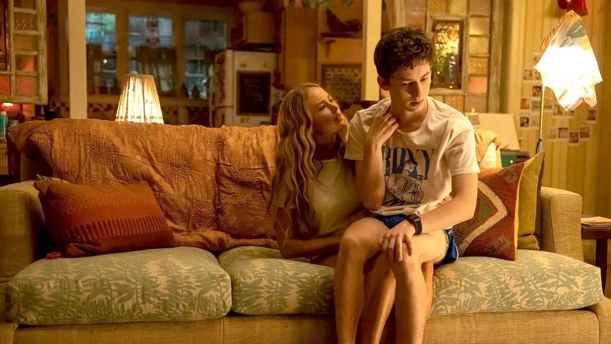 Jennifer Lawrence and Andrew Barth Feldman in a scene from 'No Hard Feelings.' Lawrence is a white woman with long blond hair wearing a white t-shirt and shorts. Feldman is a young white man with short dark hair wearing a white t-shirt and shorts. They are seated on a couch in a living room, and he is sitting on her lap looking away from her.