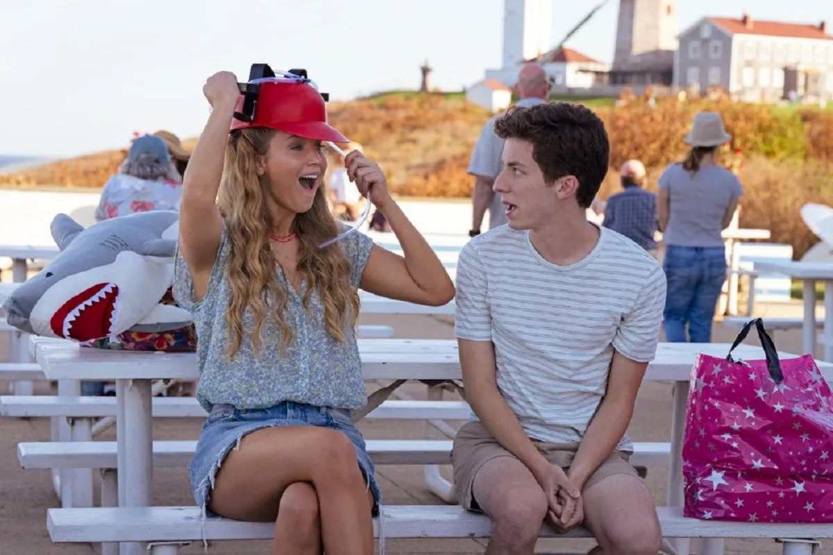 Jennifer Lawrence and Andrew Barth Feldman in a scene from 'No Hard Feelings.' They are seated at a white picnic table on the same bench at a beachside cafe. Lawrence is a white woman with long blonde hair wearing a blue patterned button-up blouse and a short denim skirt. She's putting on a red novelty baseball hat with an attachment for two beer cans on it. Feldman is a young white man with short dark hair wearing a grey and white striped t-shirt and khaki shorts. 