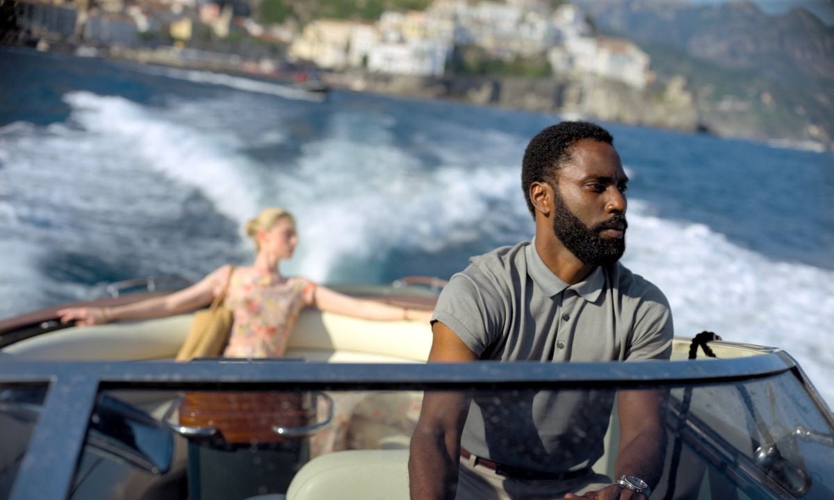 John David Washington as The Protagonist in a scene from 'Tenet.' He is a Black man with short hair and a beard wearing a grey polo shirt driving a speedboat. There is a blonde, white woman in a summer dress sitting casually in the back of the boat. 