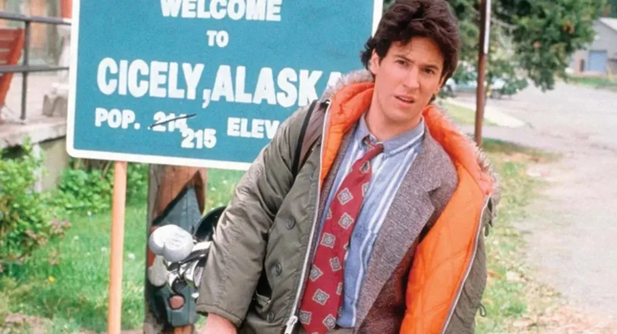 Rob Morrow in Northern Exposure; image depicts a white man with brown hair in a winter coat and suit, standing in from of sign for Cecily, Alaska