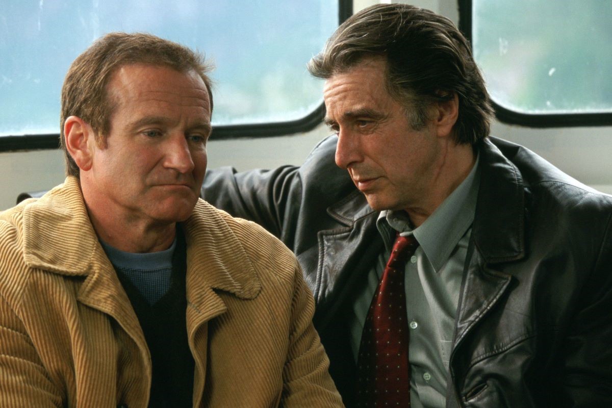 Robin Williams as Walter and Al Pacino as Will in a scene from 'Insomnia.' Walter is a white man with brown hair wearing a light brown corduroy  jacket over a blue shirt. Will is a white man with dark hair wearing a black leather jacket over a blue buttondown shirt and a red tie. They are seated closely together on a bench on a ferry. 