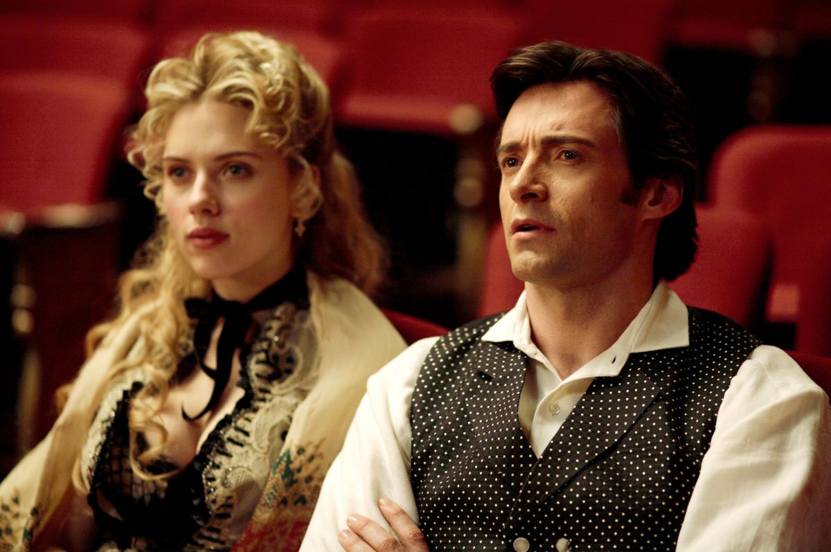 Scarlett Johansson as Olivia and Hugh Jackman as Robert in a scene from 'The Prestige.' Olivia is a white woman with long, curly blonde hair wearing a Victorian style black and white dress. Robert is a white man with dark hair wearing a white shirt under a black, Victorian-style vest. They are seated in a theater looking at something in front of them and surrounded by empty red seats. 
