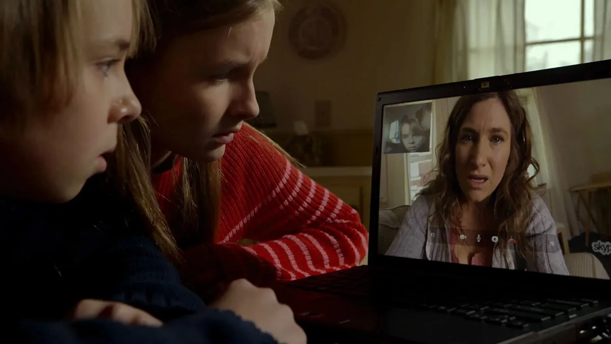 two kids hunch over a laptop with their mom's (Kathryn Hahn) face on it in The Visit