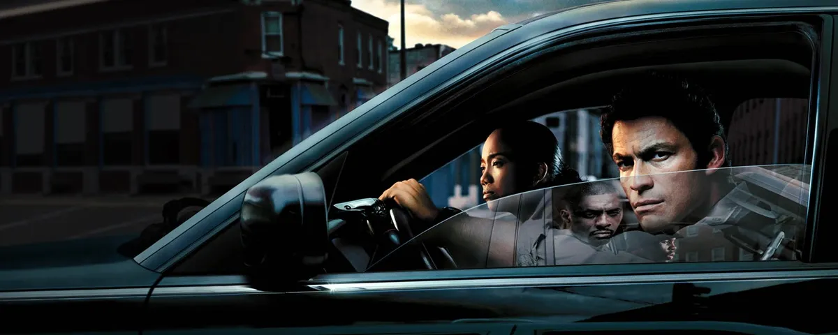 A man glares from a car window and a woman sits in the passenger seat in The wire
