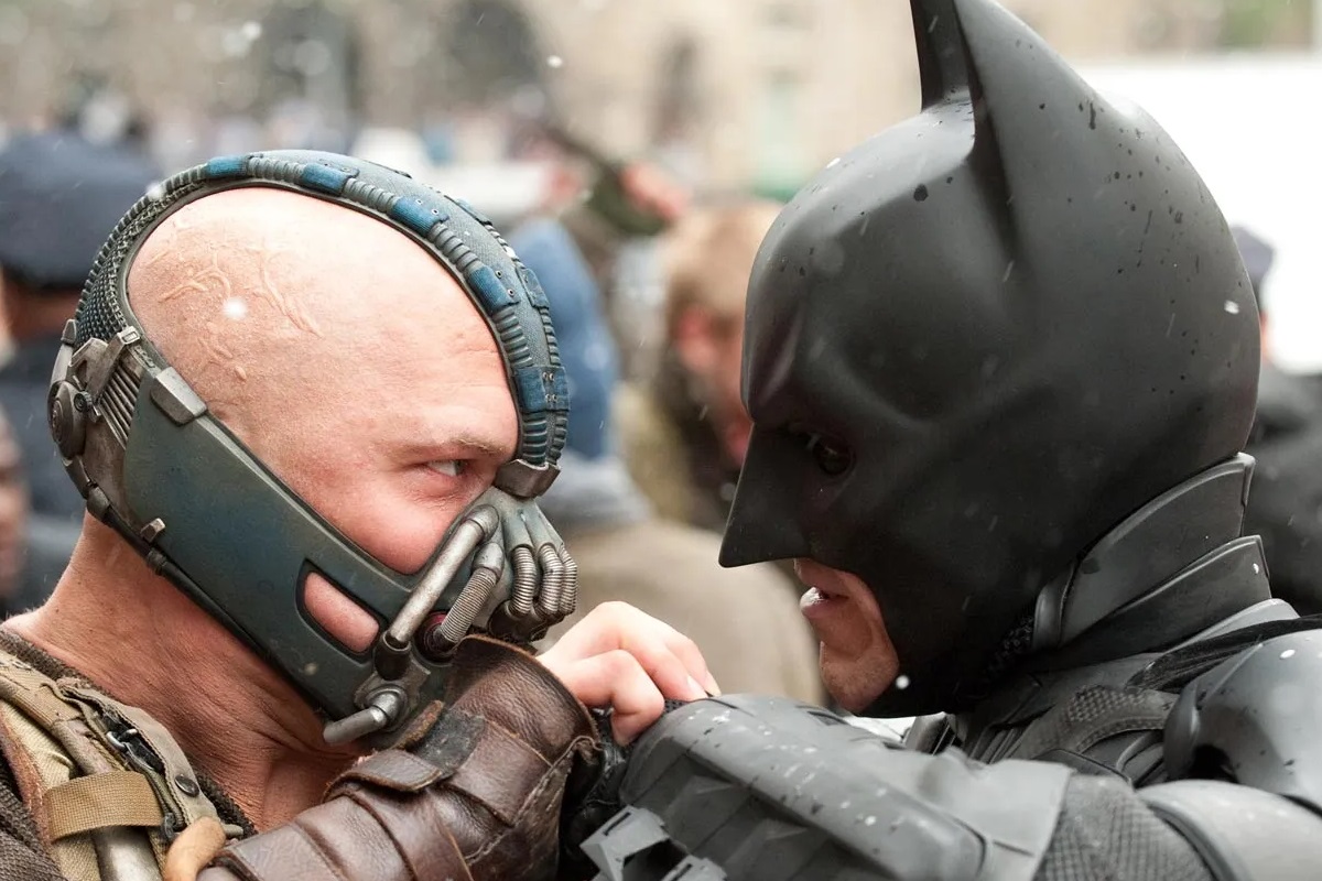 Tom Hardy as Bane and Christian Bale as Batman in a scene from 'The Dark Knight Rises.' Bane is a white man who is bald and wearing a metal mask clamped over the top of his head, and over his cheeks, nose and mouth. Batman is wearing his signature cowl over his head, revealing only his mouth. they are grappling mid-fight. 