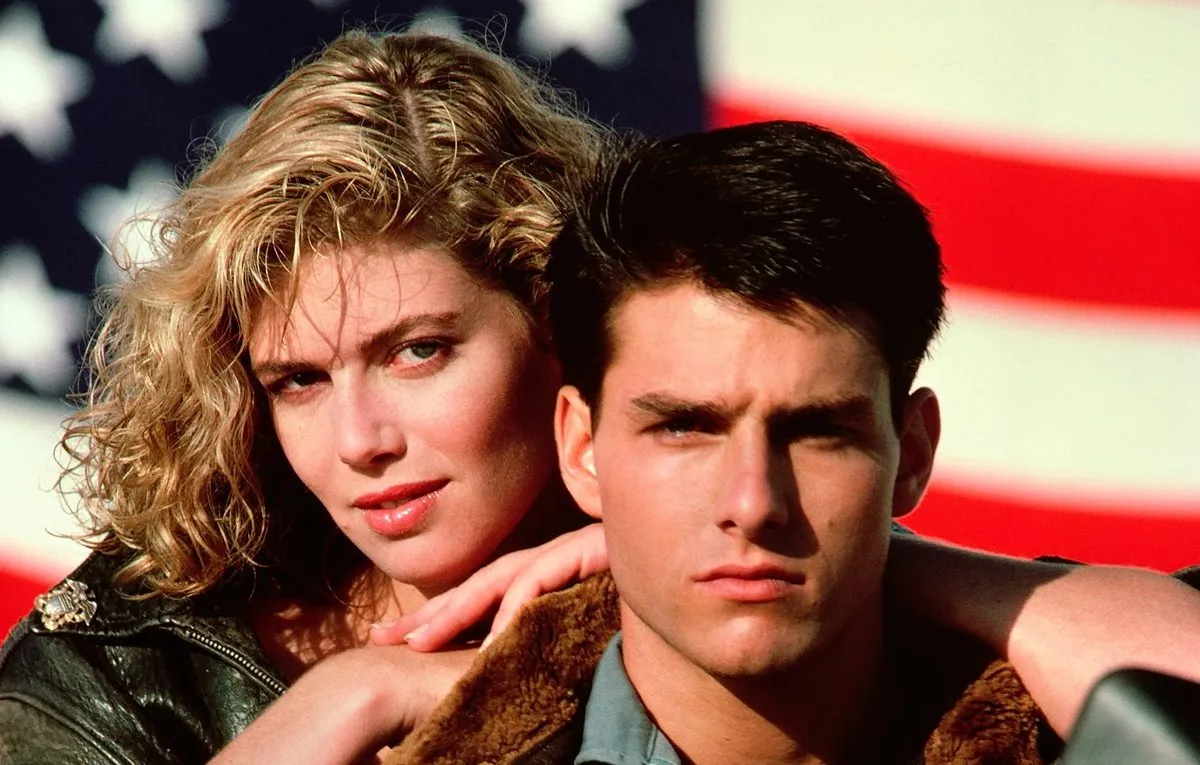 Kelly McGinnis leans over Tom Cruise's shoulder with American flag in background for Top Gun