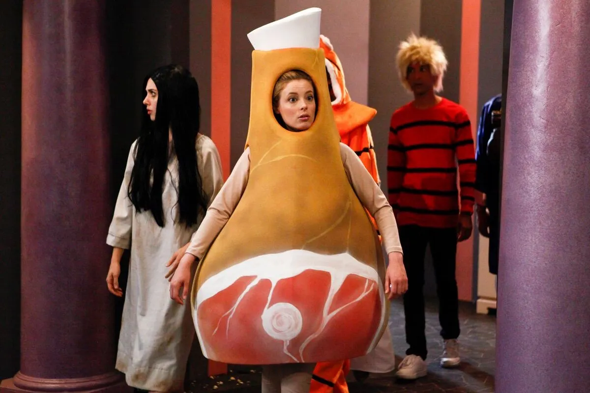 Still from Community episode Paranormal Parentage; Alison Brie as Samara from the Ring, in a white dress and long black wig, Gillian Jacobs in a ham bone suit, and Danny Pudi behind them in a scruffy blonde wig and orange and black striped top.