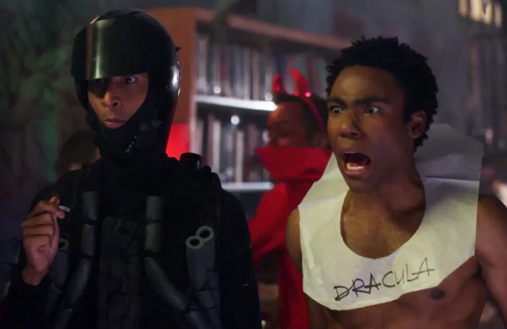 Still from Community episode Epidemiology; Danny Pudi, dressed all in black with a motorcycle helmet and tubes on his torse, next to Donald Glover, shirtless and with a toilet paper collar with Dracula written on it. Donald Glover is screaming.