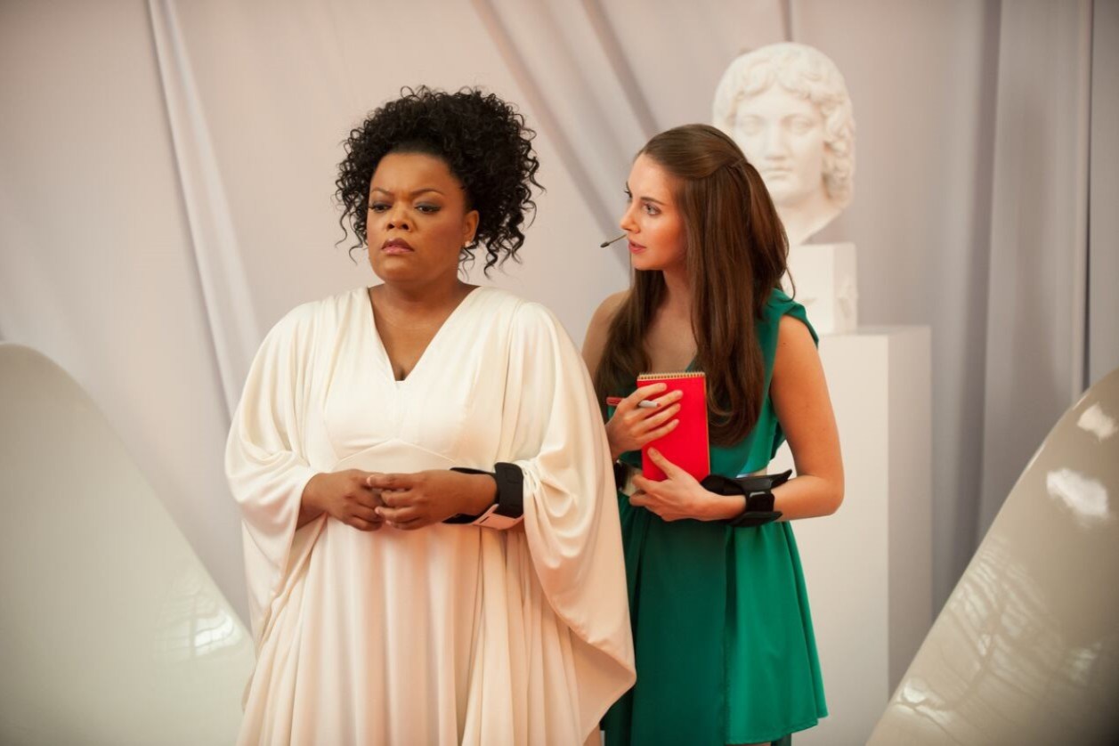 Still from Community episode App Development and Condiments; Yvette Nicole Brown stands with her hands together wearing a white drapes sleeve Grecian gown while Alison Bree in a green sci-fi Grecian tunic stands behind and to the right of her looking attentive.