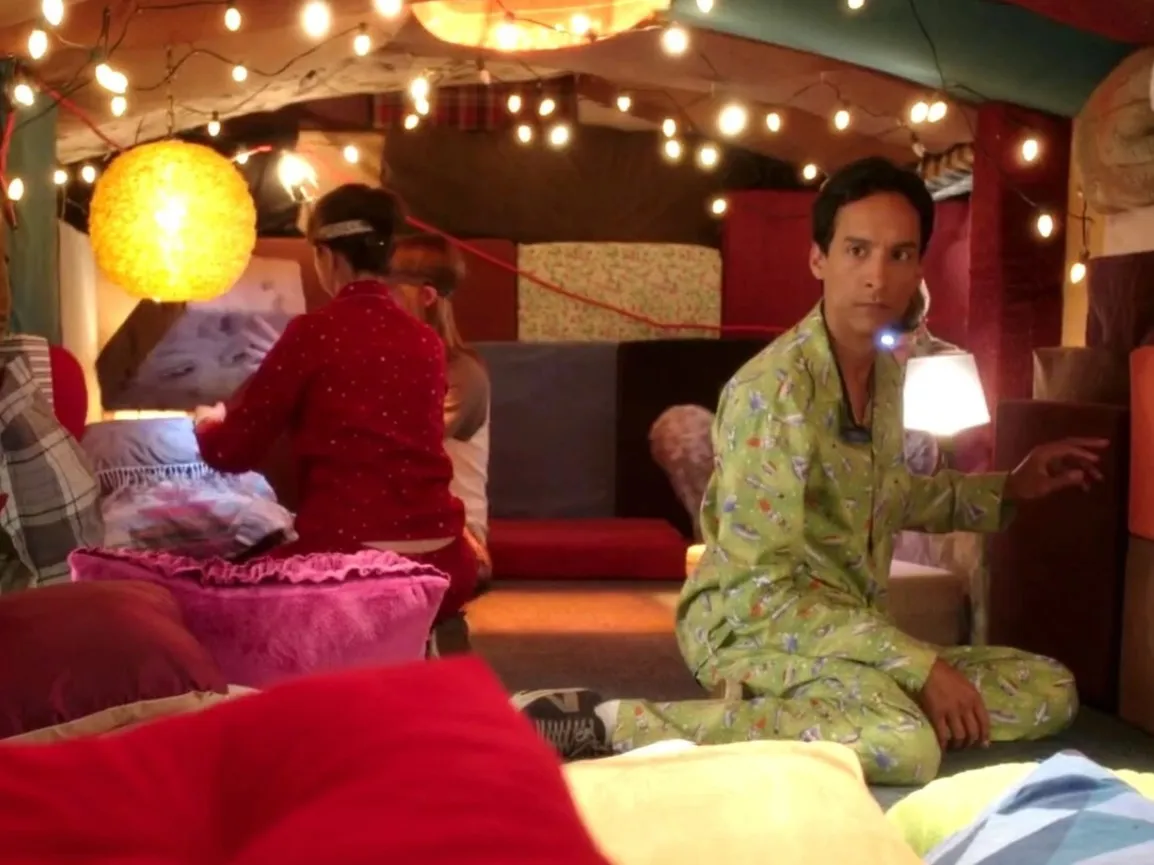 Still from Community episode Pillows and Blankets; Danny Pudi kneels inside a blanket fort wearing green pjamas. There's fairy lights over head and more people behind him.